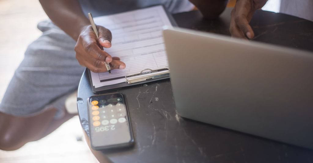 Person tallying numbers on a clipboard beside a laptop and calculator